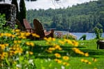 A beautifully landscaped residential lawn in Lake Geneva featuring vibrant yellow wildflowers in the foreground, three wooden Adirondack chairs facing a scenic lake, and a lush forested hill in the background.