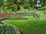 A meticulously landscaped public park or commercial garden in Pleasant Prairie featuring curved flower beds with red, white, and purple blooms, a green grass lawn, park benches, and lush green trees.